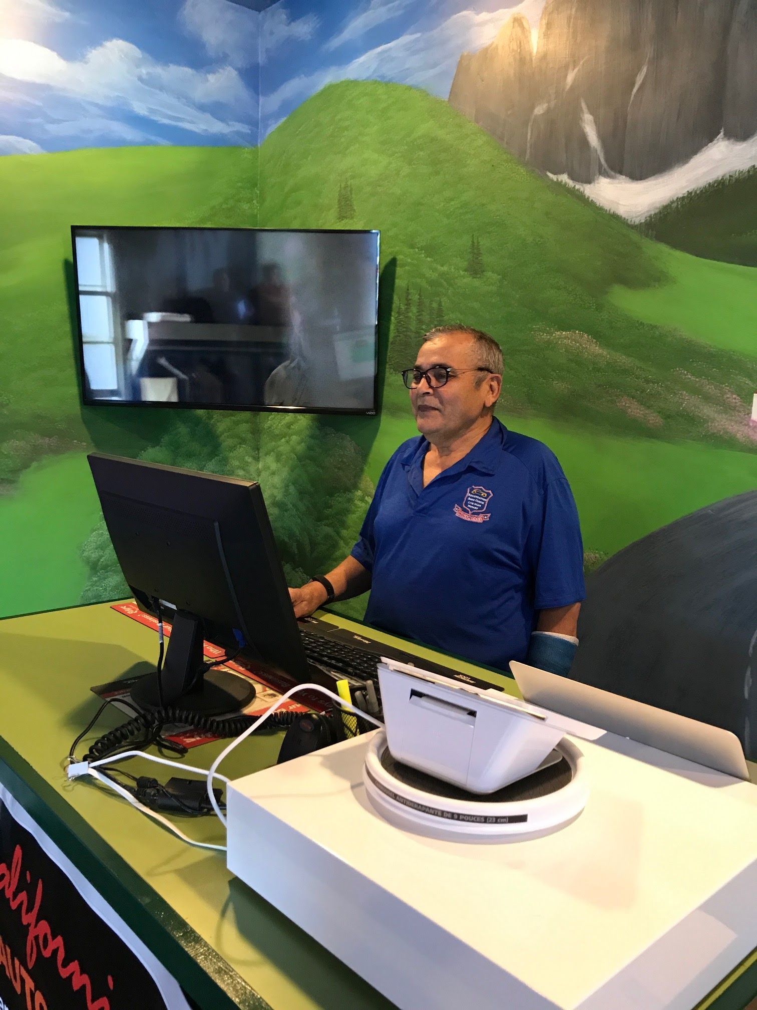 Man at a booth with a laptop and monitor in front of a green mountain mural.