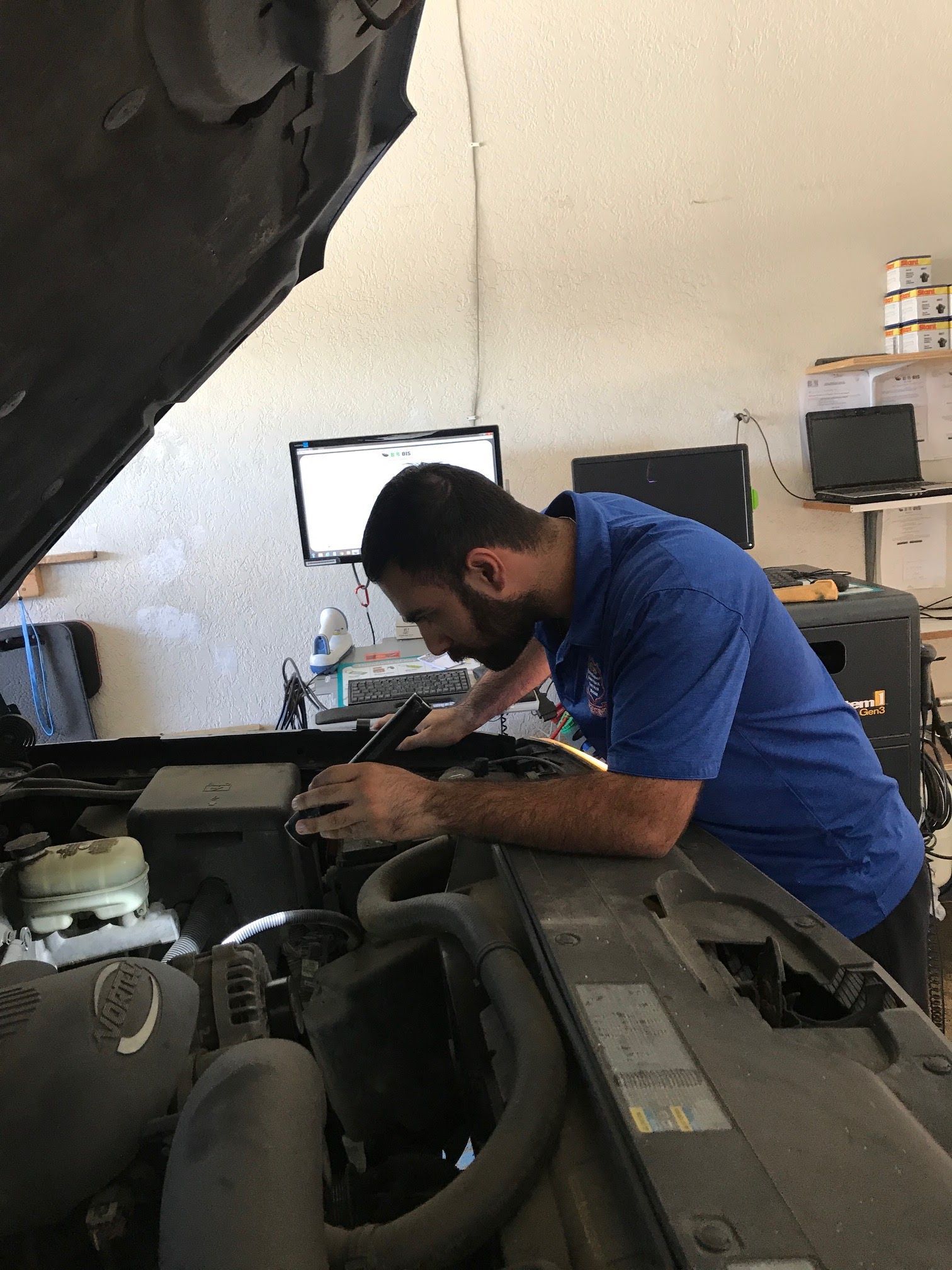 Mechanic in blue shirt working under an open car hood in a garage.
