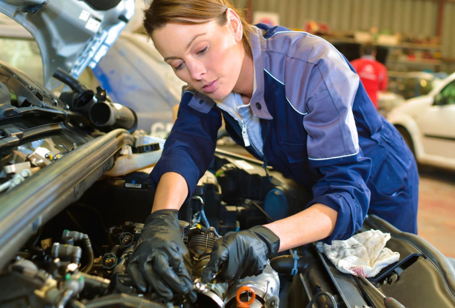 Mechanic in blue coveralls and gloves working under a car hood in a garage