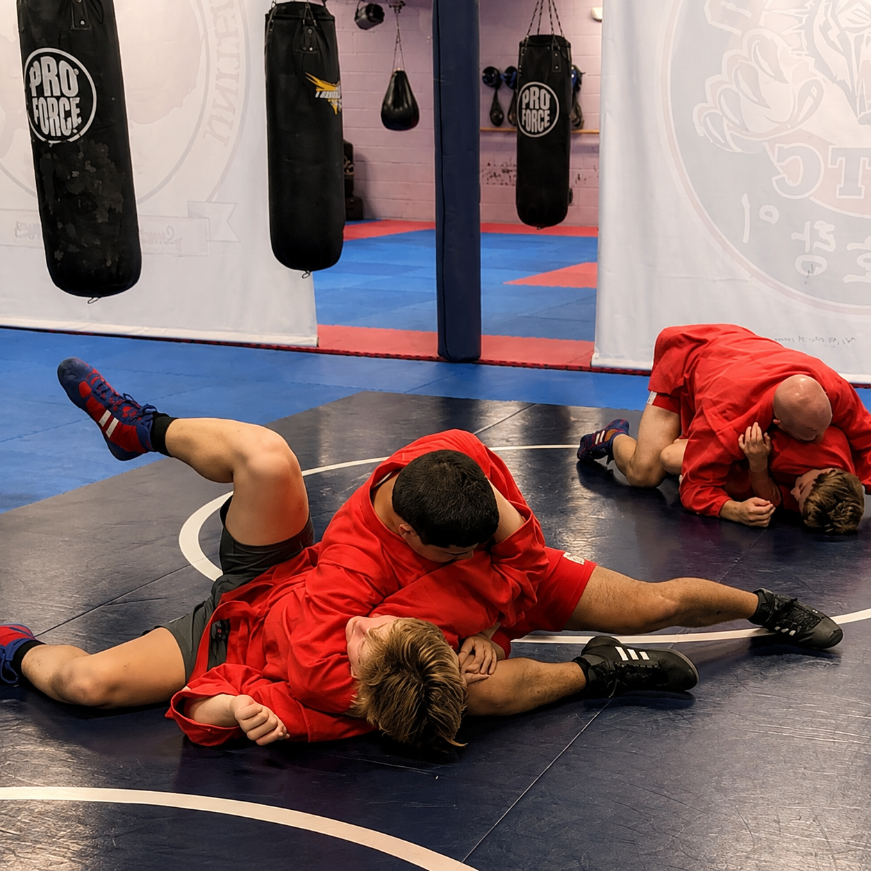 Two martial artists in red practice grappling on a blue mat in a gym with hanging punching bags.
