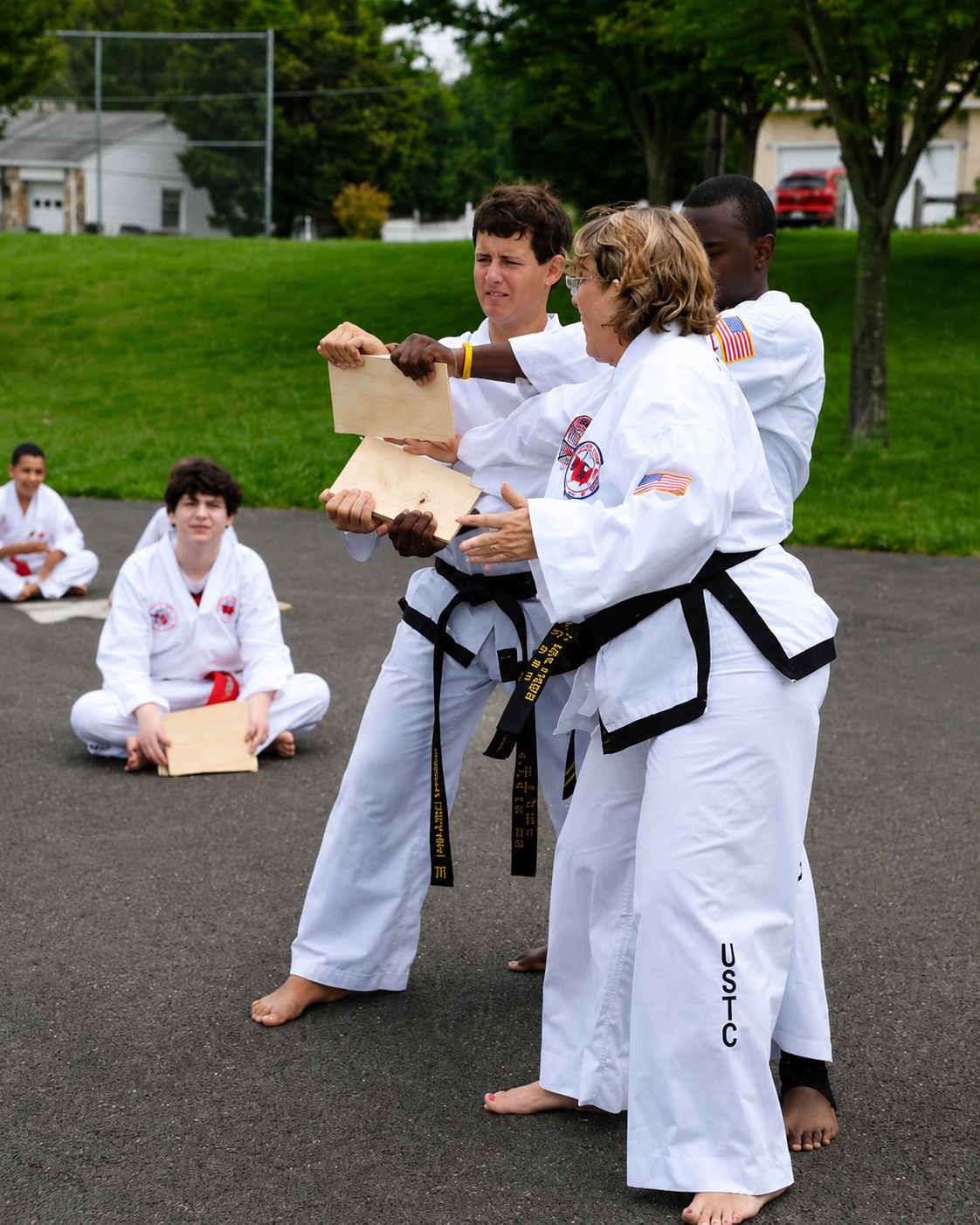 Two martial artists in white uniforms break a wooden board outdoors while others watch nearby.