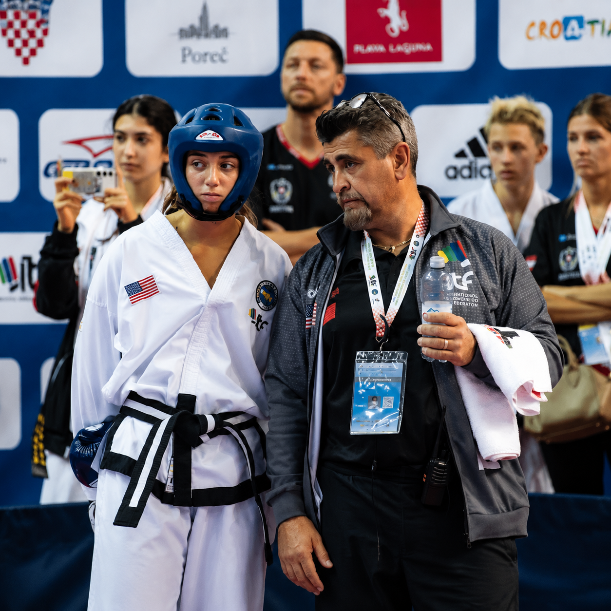 Martial arts competitor in white uniform and blue headgear stands beside coach at an awards podium
