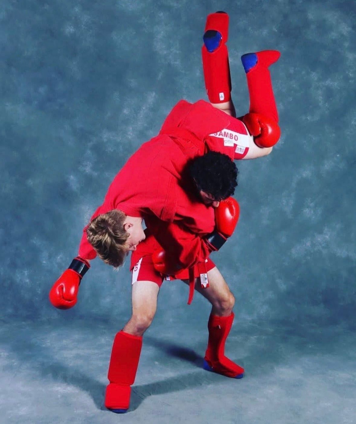 Two martial artists in red practice grappling on a blue mat in a gym with hanging punching bags.