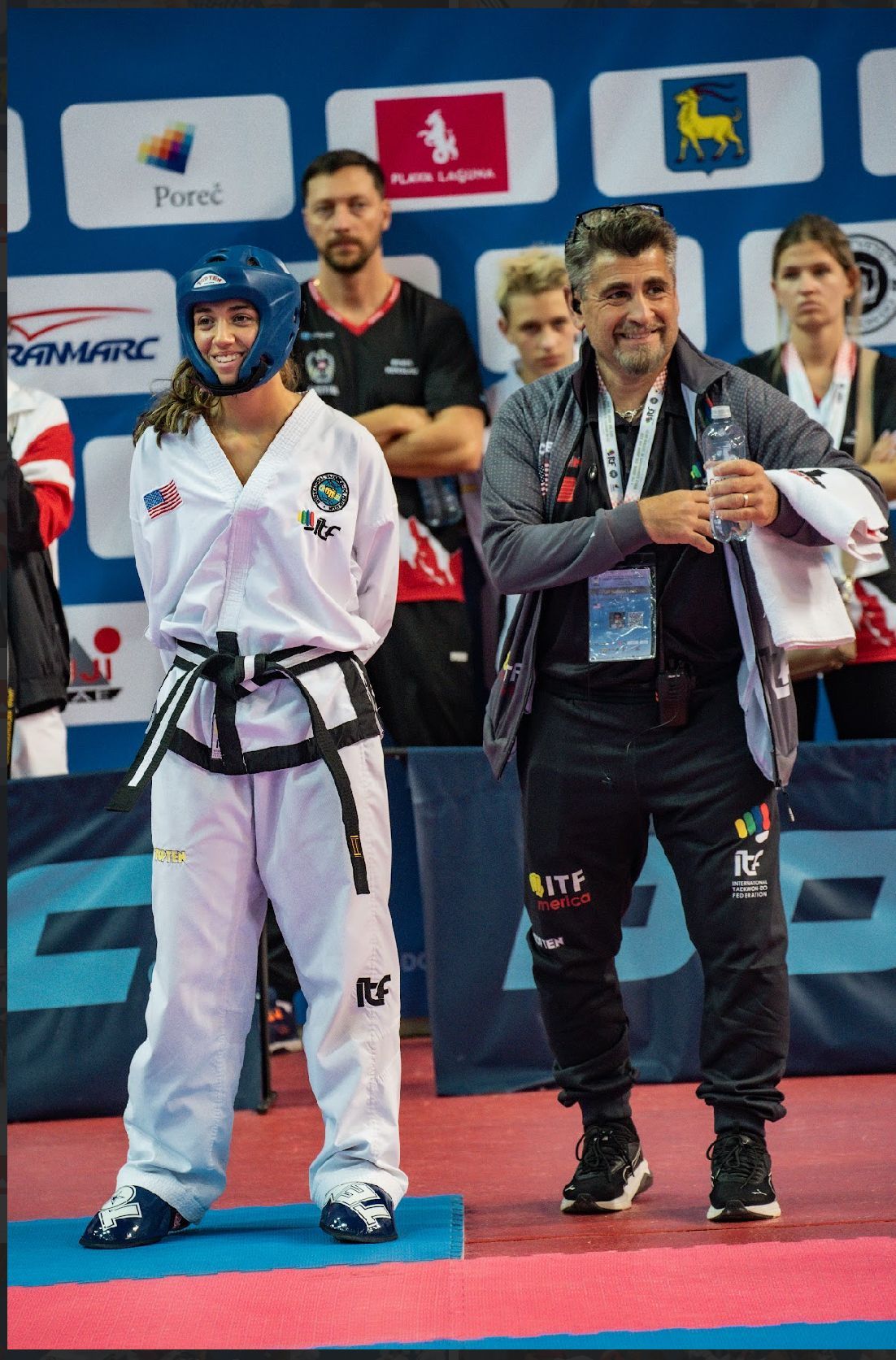 Two martial artists in white uniforms break a wooden board outdoors while others watch nearby.