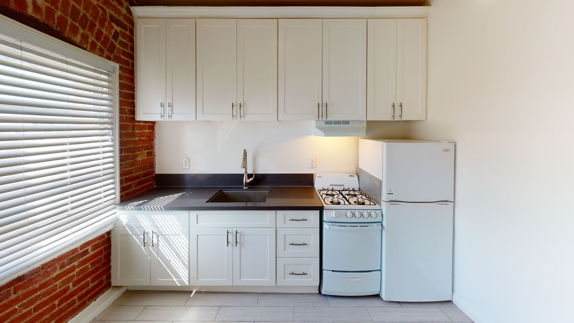 Small kitchen with white cabinets, dark countertop, gas stove, and refrigerator. Red brick wall.