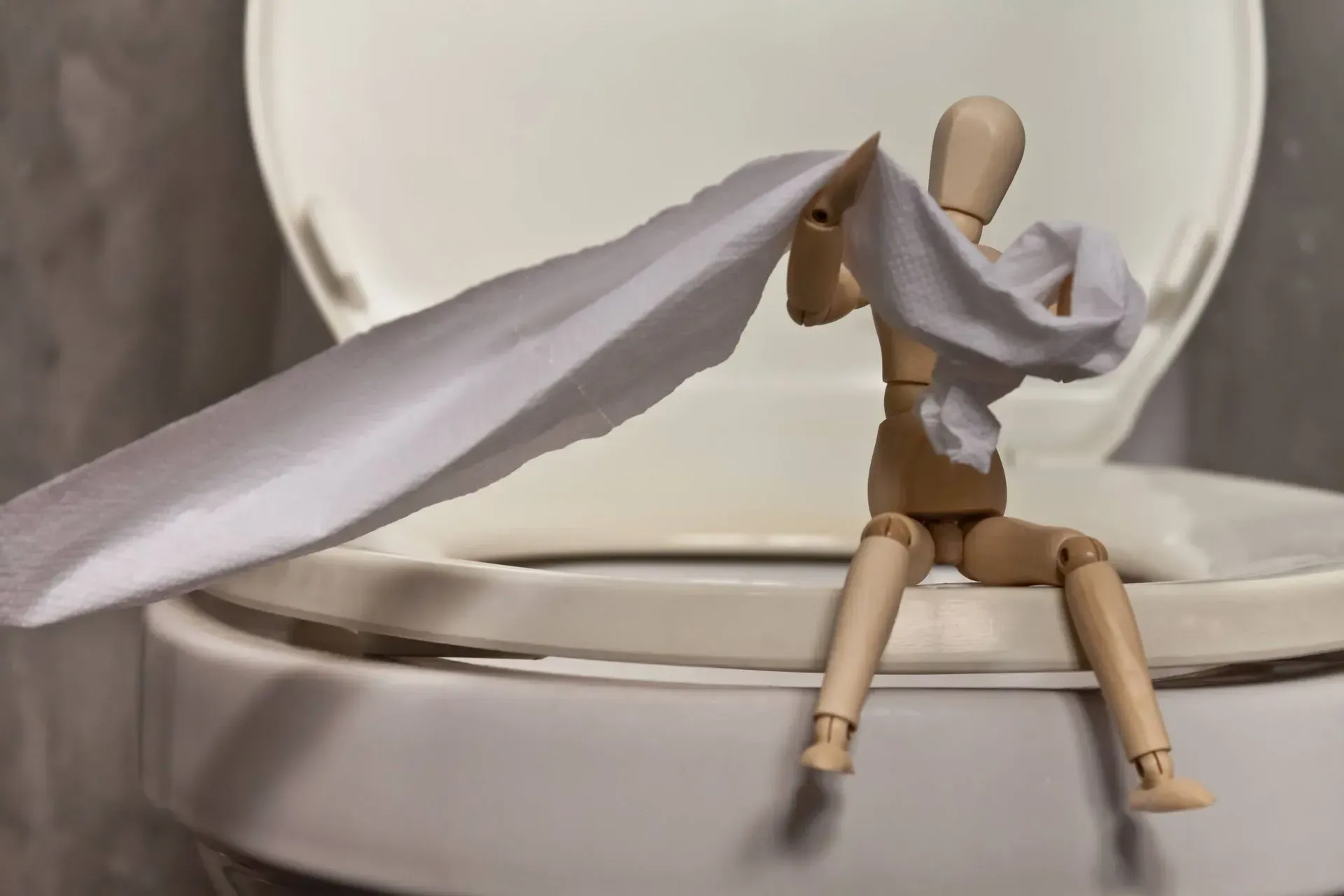 A wooden mannequin is sitting on top of a toilet holding a piece of toilet paper.