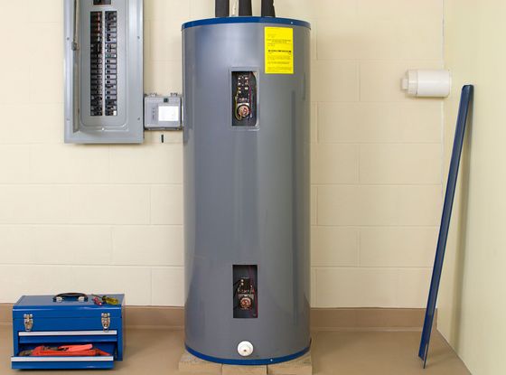 A gray electric water heater stands next to an open electrical panel, with a blue toolbox and trim piece nearby.