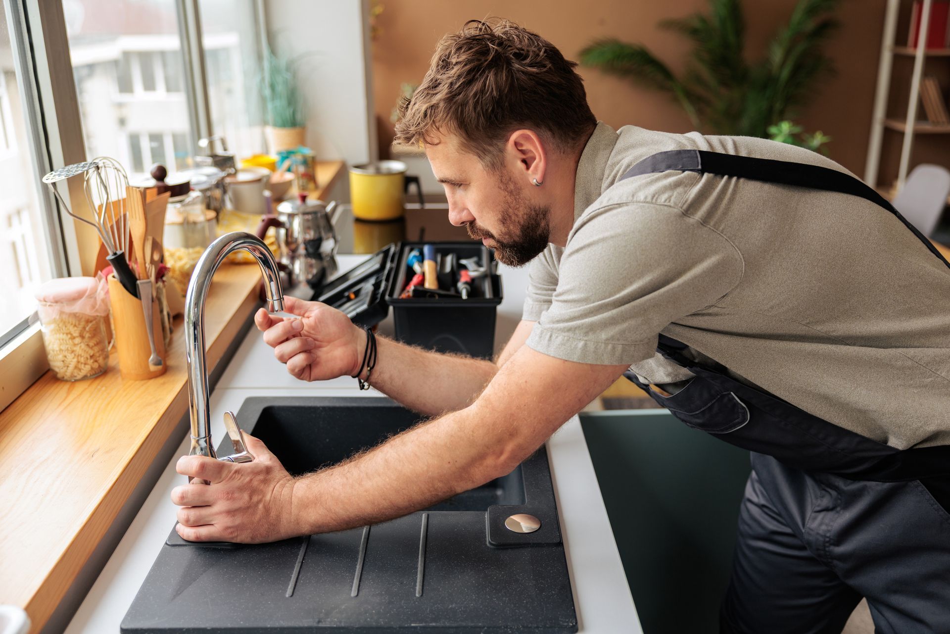 A plumber in work overalls adjusting a kitchen faucet with a toolbox nearby.