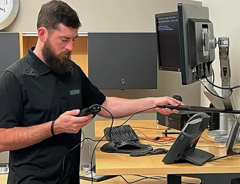 A man with a beard is working on a computer while holding a cell phone.