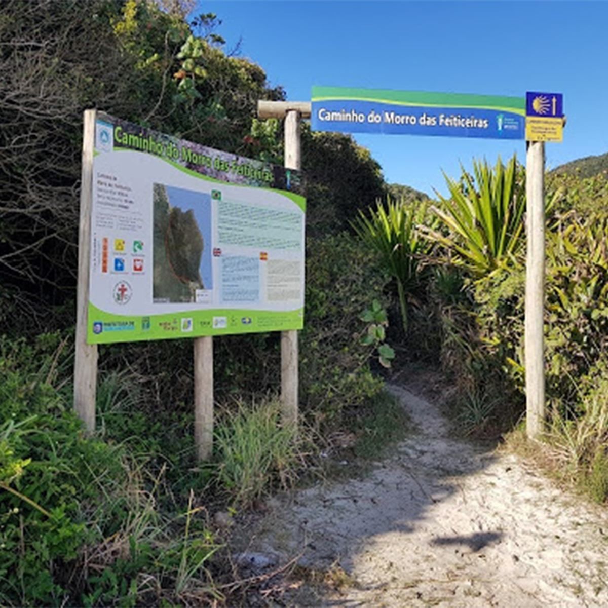 Placa de entrada da trilha ao lado de um caminho de areia em meio à vegetação costeira verdejante sob um céu azul.