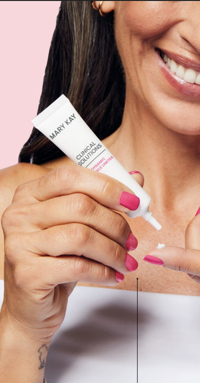 Mujer sonriendo mientras se aplica crema facial blanca de un tubo en la mano sobre un fondo rosa.
