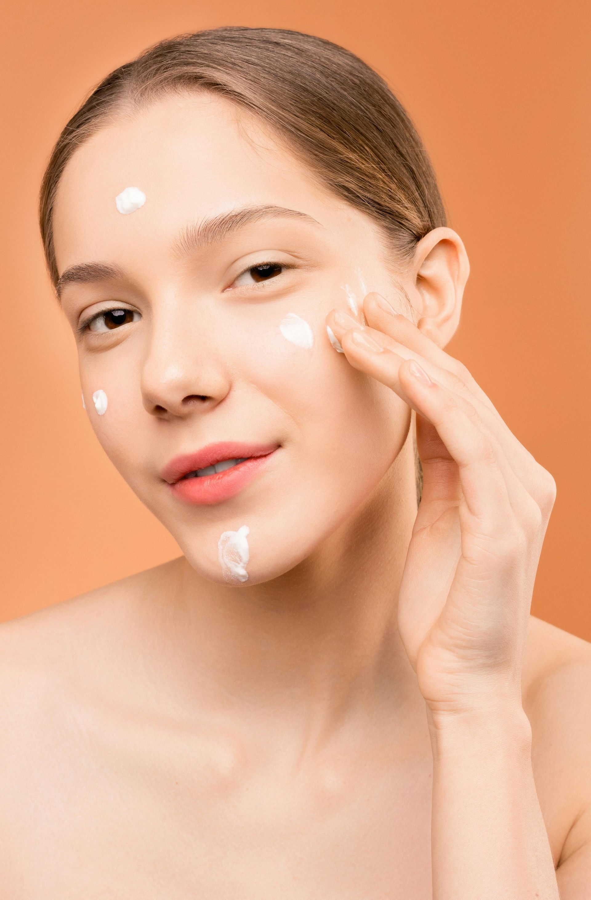 Mujer aplicándose crema en la cara sobre un fondo color melocotón.