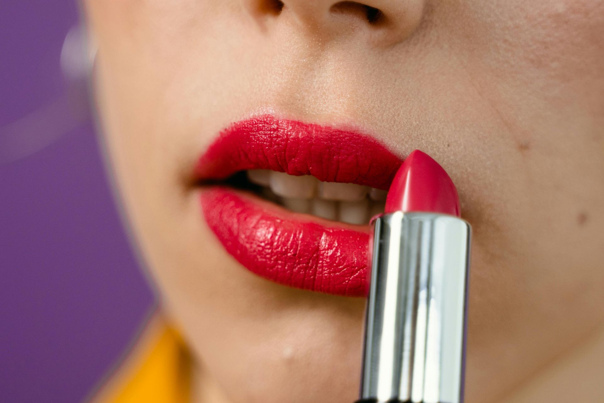 Primer plano de unos labios aplicándose un pintalabios rojo brillante con un tubo plateado sobre un fondo morado.