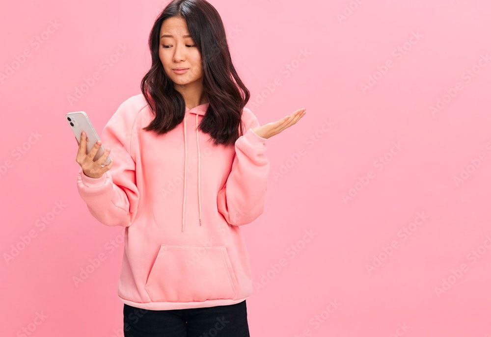 a woman in a pink hoodie is holding a cell phone and shrugging her shoulders .