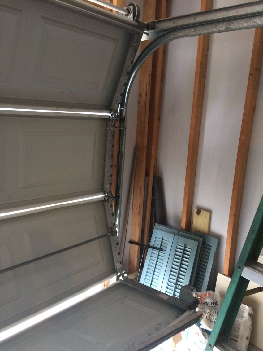 Inside a garage, a partially open metal garage door with storage items leaning against the wall.