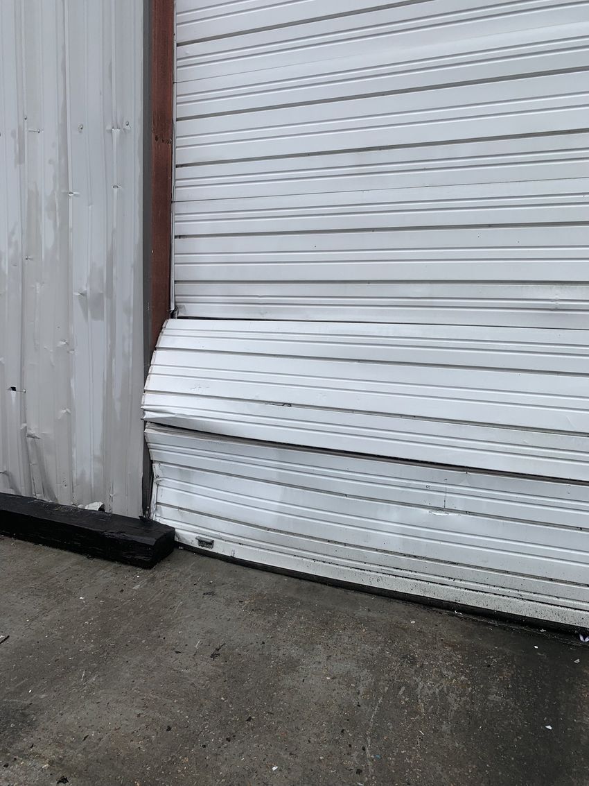 White metal roll-up door with a bent lower panel beside a gray wall on a concrete floor.
