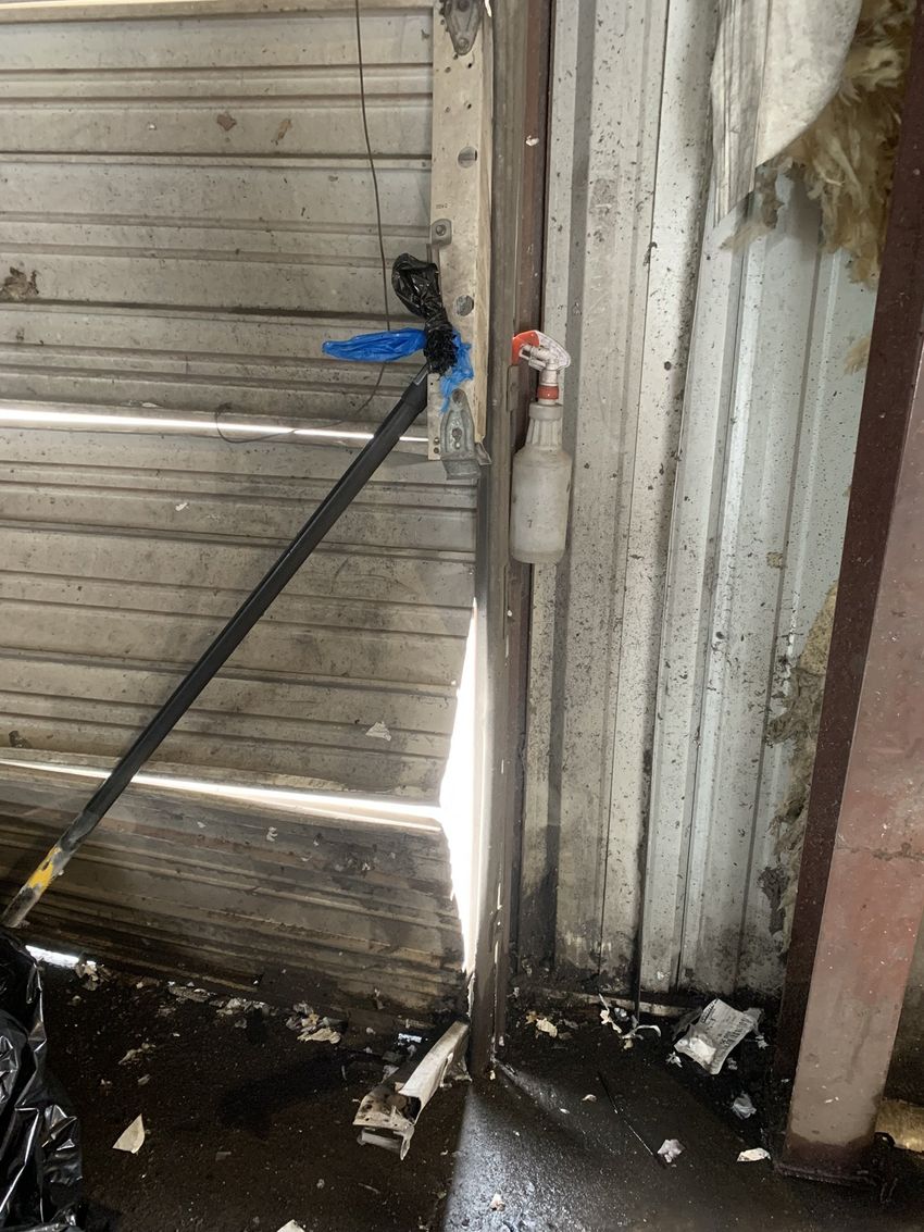 Black hose with blue clamp beside a weathered wooden gate and corrugated metal wall, with water on the ground