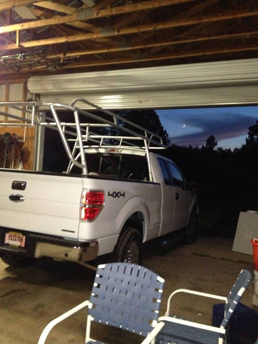 White pickup truck with ladder rack parked under a carport at dusk, with patio chairs in the foreground