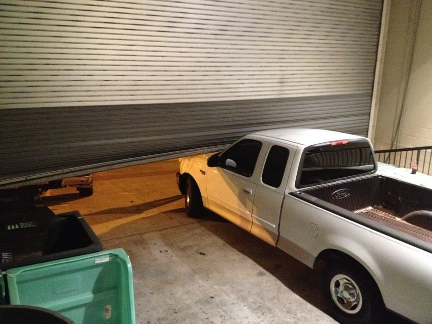 White pickup truck and green dumpster parked beside a closed garage door in a cluttered alley