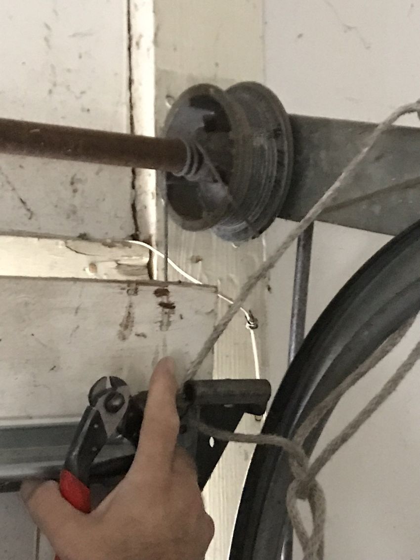 Hand using pliers to work on a cable or rod near a wall-mounted pulley or spool.