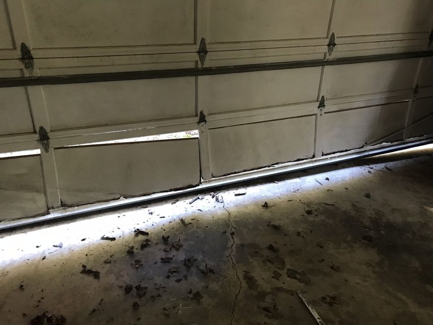 Bottom of a garage door with a gap, showing dirt and debris on the floor.
