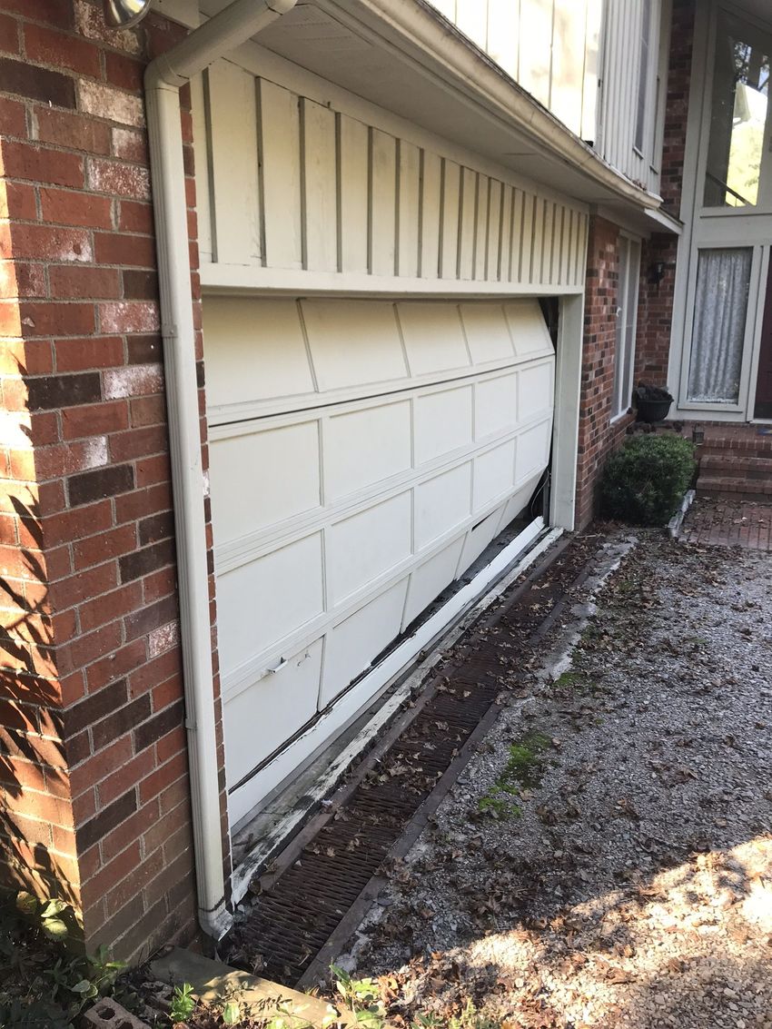 Damaged white garage door sagging open beside a brick house and gravel driveway