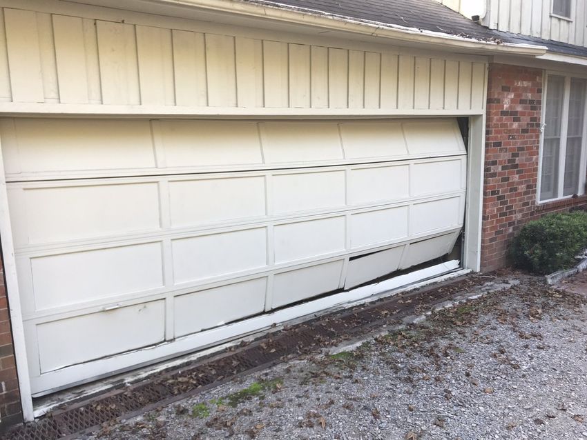 White garage door with a dented lower panel beside a brick wall and gravel driveway