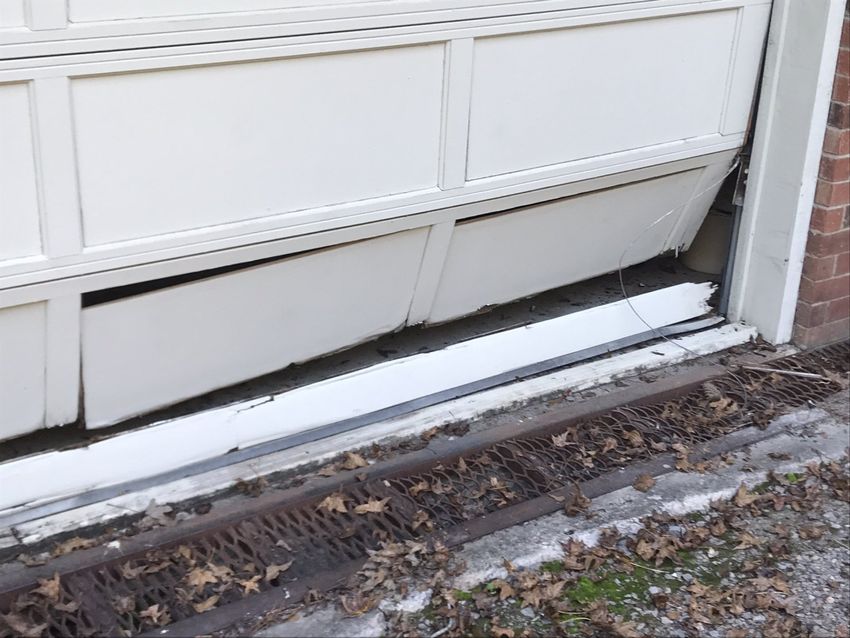 Broken white garage door with snow on the bottom edge and a gap along the driveway side