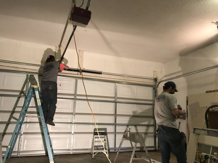 Two people using a ladder and pole tool to install or adjust equipment in a garage.