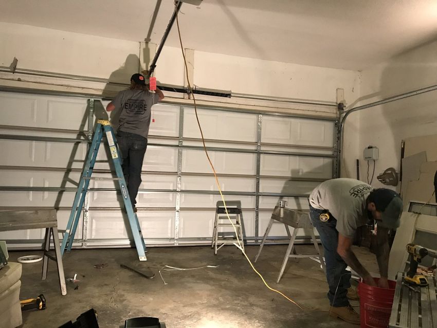 Two workers installing a garage door track, one on a ladder and one bending near tools.