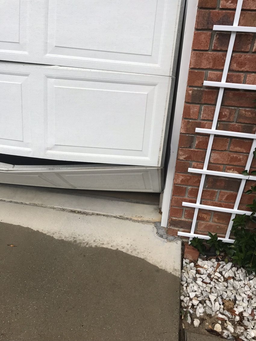 White garage door with cracked concrete threshold beside a brick wall and white stone landscaping.