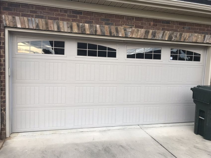 White two-car garage door on a brick house, with a driveway and trash bin visible.