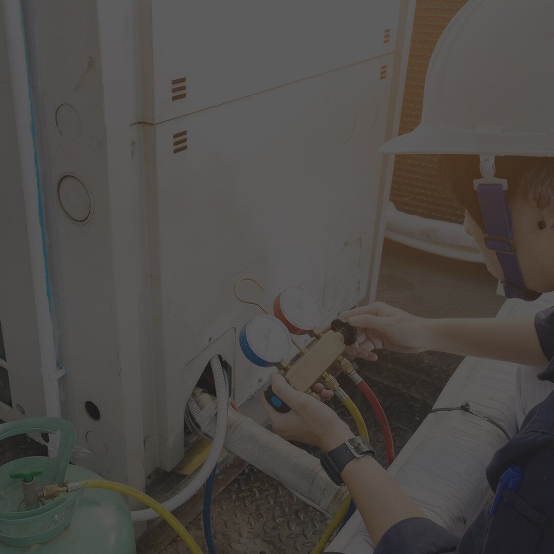 An HVAC technician wearing a hard hat is servicing an outdoor unit with gauges and hoses.