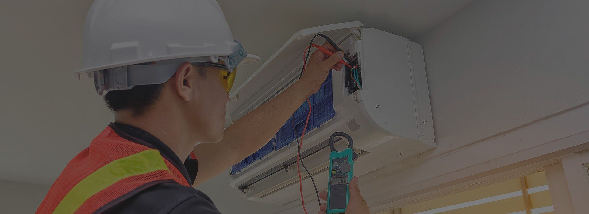 An electrician wearing a hard hat and safety vest is inspecting an air conditioning unit attached to a wall.