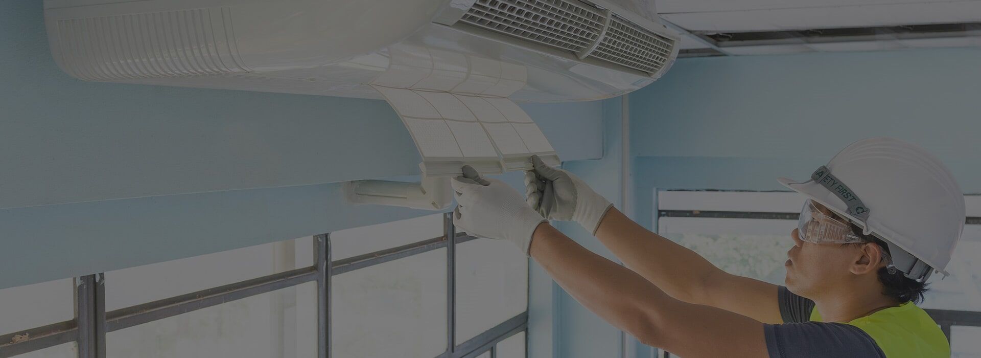A person in a hard hat and gloves is servicing an air conditioning unit in a room with blue walls.