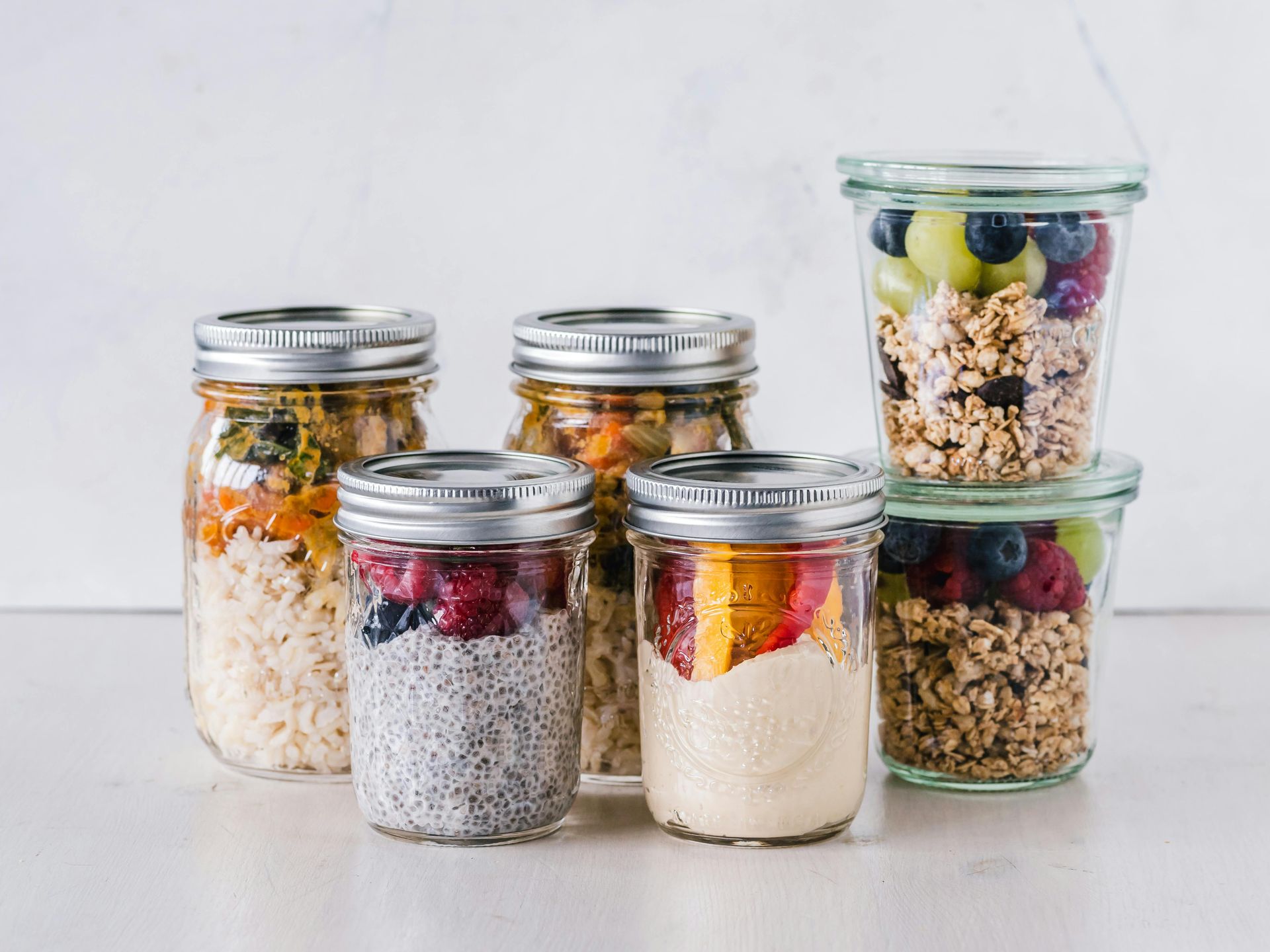 Frascos de vidrio llenos de alimentos en capas, como granola, fruta y pudín de semillas de chía, sobre una superficie blanca.