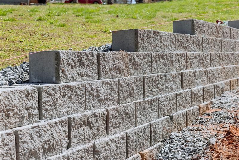 A tiered retaining wall made of gray concrete blocks set against a grassy slope with gravel drainage at its base.