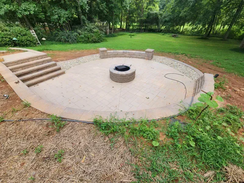 A circular stone patio with a central fire pit, curved seating wall, and stone steps, set in a backyard near woods.