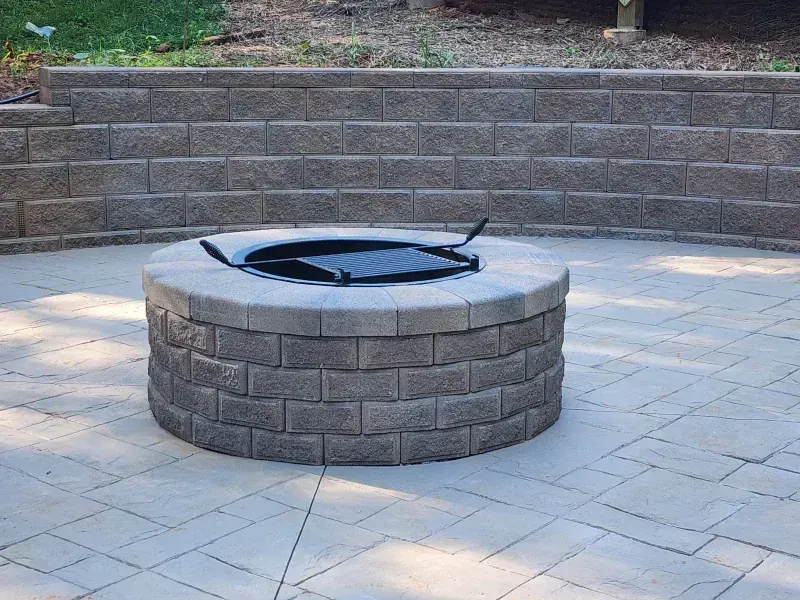 A circular stone fire pit with a metal cooking grate sits on a paved patio in front of a curved stone retaining wall.