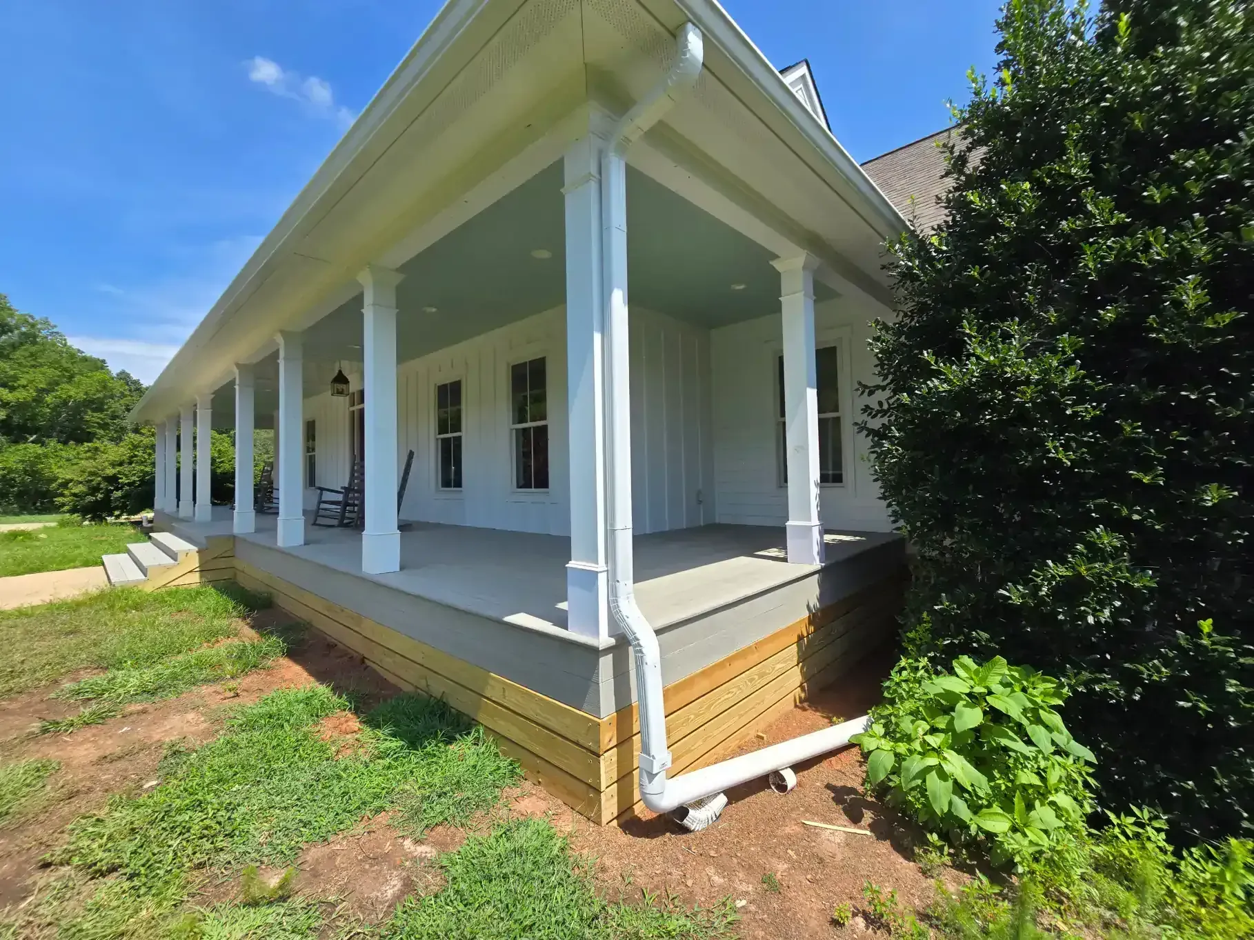 A white farmhouse with a large, covered front porch, white columns, a blue porch ceiling, and natural wood skirting.