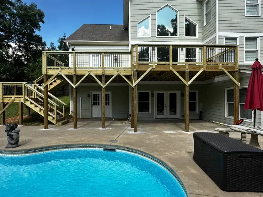 A multi-level wooden deck with a staircase overlooks a backyard swimming pool and a light gray house.