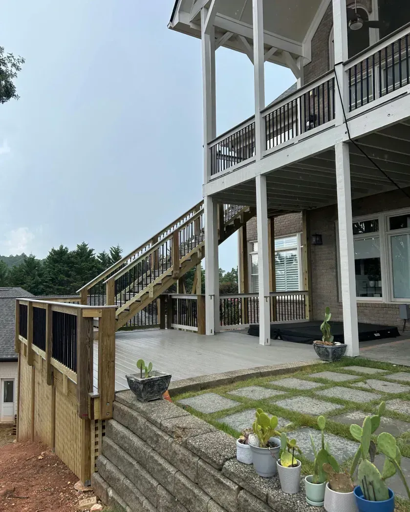Wooden deck with stairs descending to a paved patio area at the base of a multi-story house, featuring potted cacti.