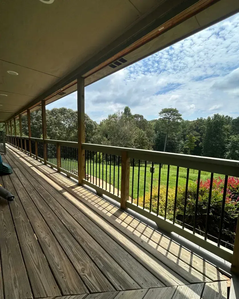 A wide wooden deck with dark metal railings overlooks a green yard and dense trees under a blue, cloudy sky.