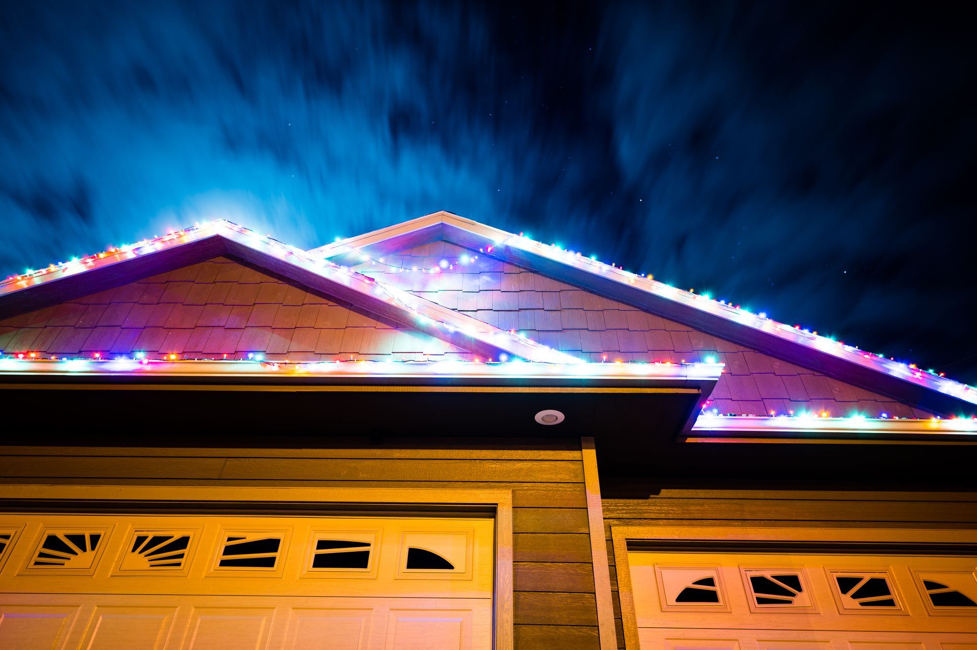 Colorful Christmas lights outline the roofline of a residential garage at night against a dark, cloudy sky.