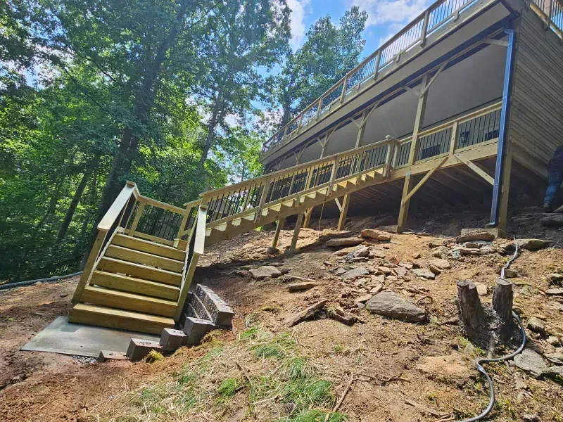A wooden staircase leads up a rocky, sloped hillside toward a large, elevated deck attached to a cabin in the woods.