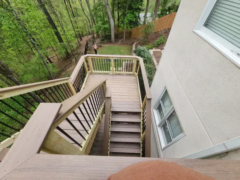A wooden deck staircase descends from a tan house toward a grassy yard and forest.