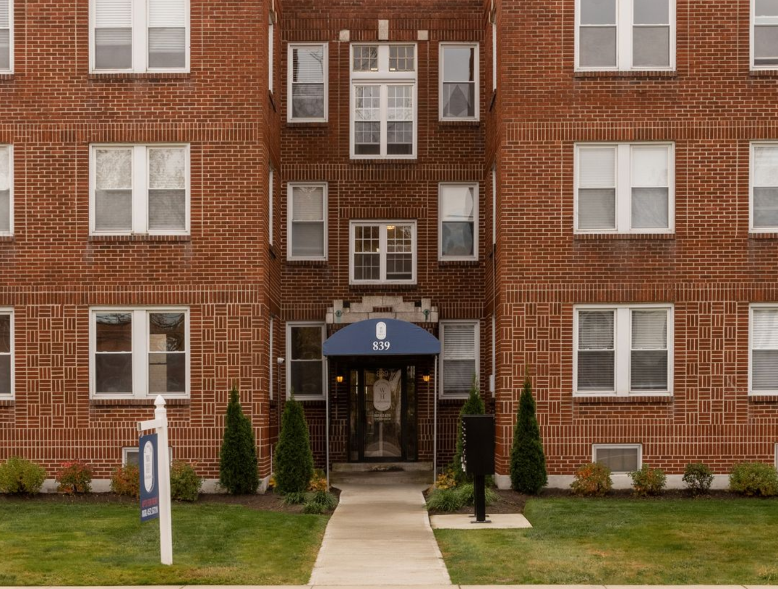 Red brick apartment building with white windows and blue awning over entrance.