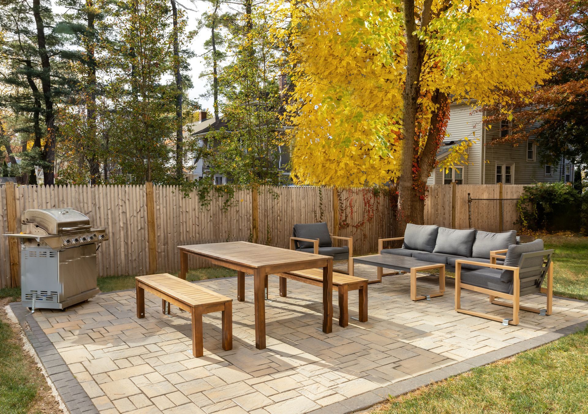 Outdoor patio with grill, dining table, benches, sofa, and chairs on a stone patio. A fence and autumn trees are in the background.