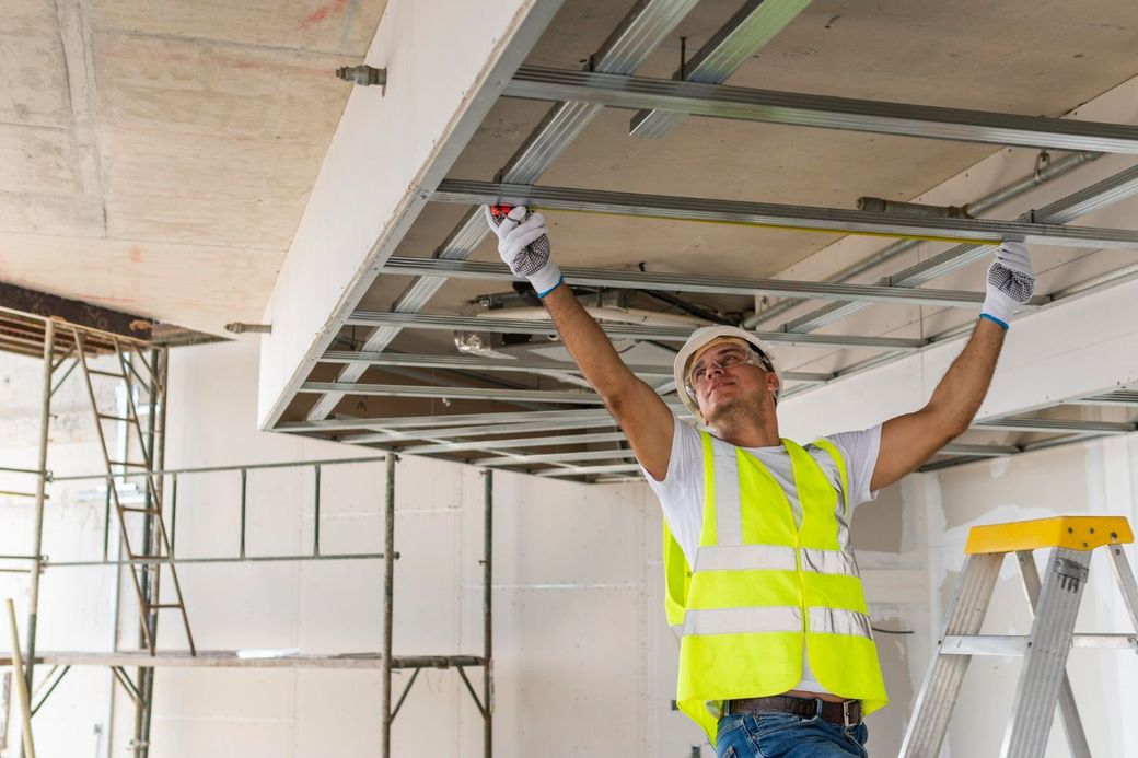 Construction worker installing metal framework on a ceiling; yellow safety vest and gloves.