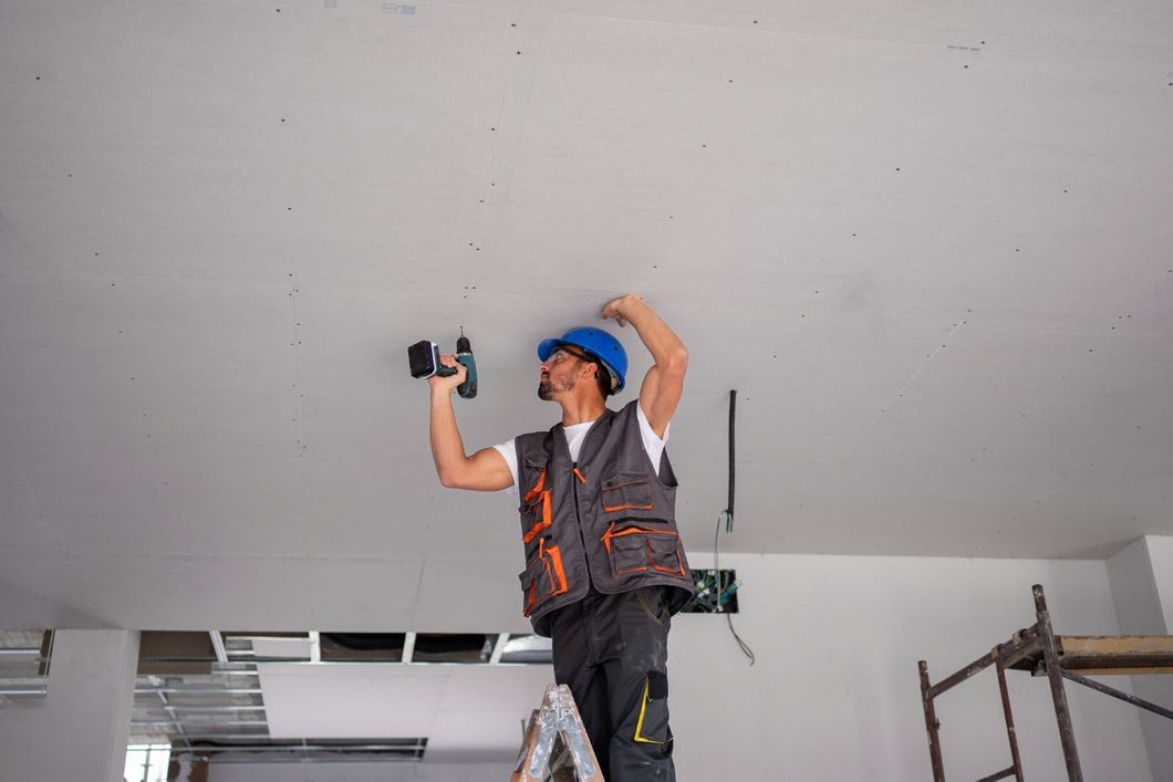 Construction worker on a ladder drills into a white ceiling. He wears a blue hard hat and safety vest.